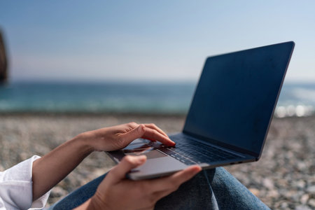 Laptop Beach Working: Woman types outdoors on laptop by sea during daytime for remote work.