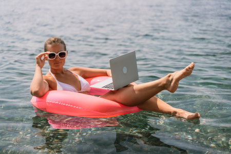A woman is floating in a pink inflatable raft with a laptop in her hand. She is smiling and she is enjoying her time in the water.の写真素材