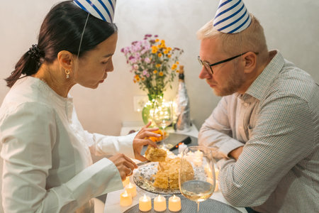 Couple Birthday Cake Celebration: Indoor intimate birthday celebration, a couple wearing party hats cuts a cake together.