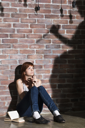 Caucasian girl sitting on the floor reading a horror or scary thriller book. Dramatic lighting with girls eyes wide open and long shadows of woman with knife on the wall near her. Terrified expressionの写真素材