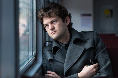 Traveling by train. Sad man traveling by train, looking through the window and thinking about unrequited love squeezing the phone in his hand. alone in an empty train wagon. Real people series.の写真素材