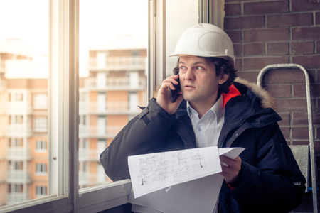 Portrait of concerned unshaven builder telling by mobile while keeping different projects in hand. Communication and work concept. Soft focus, tonedの写真素材
