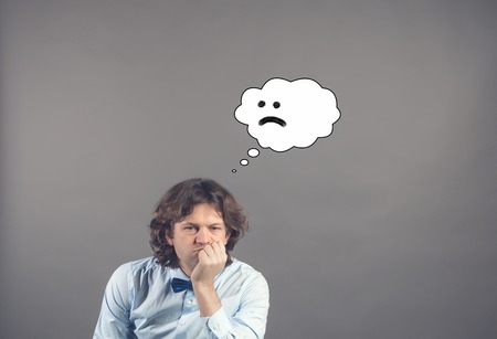 Confused upset handsome young man in blue shirt and bow tie leaned his chin on hand and looking down with drawn cloud of thoughts with sad smile in it. Grey background. Bad day. Hardship, unhappiness.の写真素材