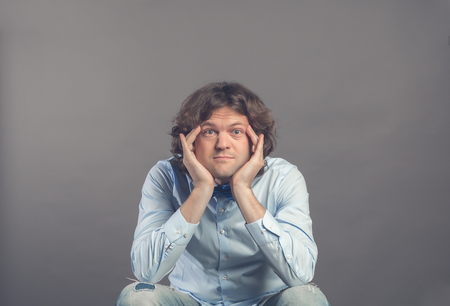 Portrait of young man in studio, resting chin on hands, while sitting and rested elbows on knees. Eyes wide open, looking at camera and smiling. Wearing blue shirt, jeans, bow tie. Dark curly hair.の写真素材
