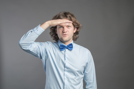 Handsome young man with dark curly hair wearing blue shirt and bow tie putting hand on his forehead and smiling, looking far isolated on grey background. Looking or waiting for someone or something.の写真素材