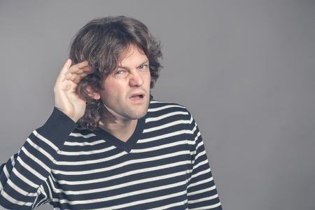 Man with impaired hearing struggling to hear frowning as he holds his hand to his ear in an attempt to improve acoustics. Gossip. Angry man holding hand to ear listening somebody. Body languageの写真素材