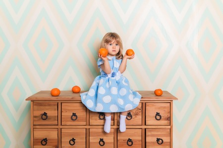 Toddler child baby girl in light blue dress seriously sitting on the wooden chest of drawers and holding fresh orange mandarins in her hands on rhomb wallpaper background. Winter holidays or christmasの写真素材