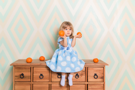 Toddler child baby girl in light blue dress seriously sitting on the wooden chest of drawers and holding fresh orange mandarins in her hands on rhomb wallpaper background. Winter holidays or christmasの写真素材