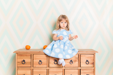 A little baby girl in her room sitting cross-legged on chest of drawers with tangerines on the rhomb wallpaper background. Child in blue polka dot dress and white socks attentively looking at cameraの写真素材
