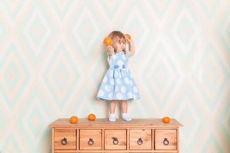 Toddler child girl in blue dress standing on wooden chest of drawers and holding fresh orange mandarins in her hands and going to throw them on rhomb wallpaper background. Winter holidays or christmasの写真素材
