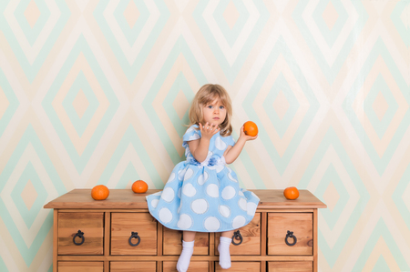 Toddler child baby girl in light blue dress sitting on the wooden chest of drawers seriously looking furrowing brows and holding fresh orange mandarins in left hand on rhomb wallpaper backgroundの写真素材