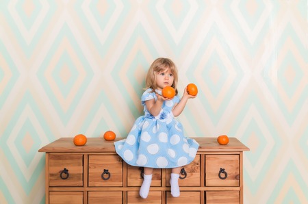Toddler child baby girl in light blue dress seriously sitting on the wooden chest of drawers and holding fresh orange mandarins in her hands on rhomb wallpaper background. Winter holidays or christmasの写真素材