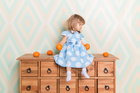 Toddler child baby girl in light blue dress seriously sitting on the wooden chest of drawers and holding fresh orange mandarins in her hands on rhomb wallpaper background. Childrens emotions feelingsの写真素材