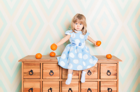 Toddler child baby girl in light blue dress seriously sitting on the wooden chest of drawers and holding fresh orange mandarins in her hands on rhomb wallpaper background. Childrens emotions feelingsの写真素材
