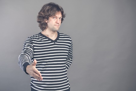 Welcome, nice to see you. Man in black and white striped sweater looking at camera with reaching out his hand offering handshake. Indoor studio shot. Hello, greeting gesture. Human emotions and signs.の写真素材