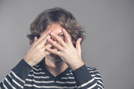 Handsome man with dark curly hair, bristle, hides face with hands, peeks through fingers, wears striped sweater, feels shy, isolated over grey background. Curious guy cannot to look at present.の写真素材