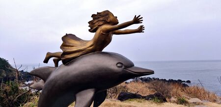 A girl statue with outstretched arms looking at the blue sky on a dolphin structure in Jeju Island, South Koreaの写真素材