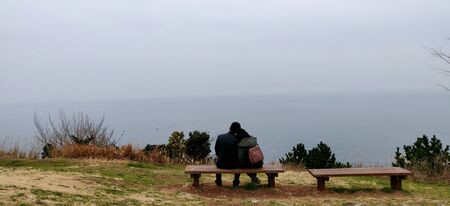 An Indian couple sitting with woman's head on man's shoulders at a bench on cliff onlooking at the vast ocean and sky during winters in jeju island, South Koreaの写真素材