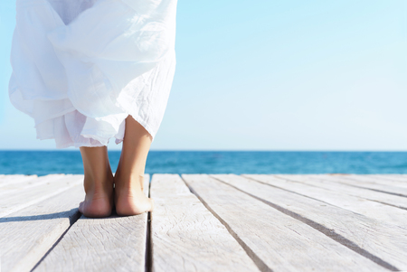 Legs of a woman in white dress on a wooden pier on summer.  Sea and sky background. Vacation, traveling and freedom concept.の写真素材