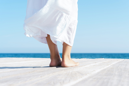 Legs of a woman in white dress on a wooden pier on summer.  Sea and sky background. Vacation, traveling and freedom concept.の写真素材