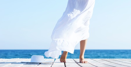 Legs of a woman in white dress on a wooden pier on summer.  Sea and sky background. Vacation, traveling and freedom concept.の写真素材
