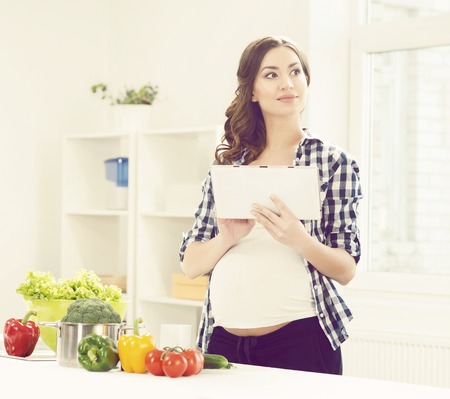 Beautiful, pregnant woman in kitchen: surfing in tablet. Motherhood, pregnancy, maternity concept.の写真素材