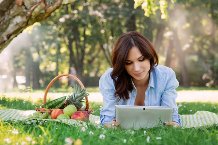 Beautiful, young brunette being addicted to the tabletduring a picnic in the park.の写真素材