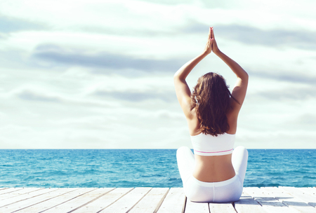 Attractive girl in white sporty clothes doing yoga on a wooden pier. Yoga, sport, leisure, recreation and freedom concept.の写真素材