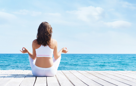 Woman in white sporty clothes meditating on a wooden pier on summer. Sea and sky background. Yoga, sport, vacation, traveling and freedomの写真素材
