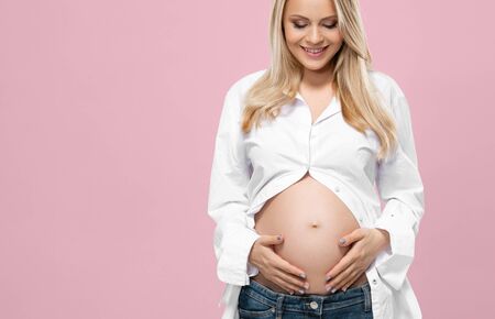 Young pregnant woman in studio embracing her beautiful tummy stomach over pink background. Pregnancy and childbearding.の写真素材