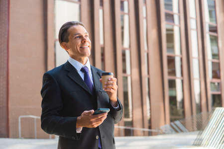 Confident businessman in front of modern office building. Financial investor is using smartphone outdoor. Banking and business.の写真素材