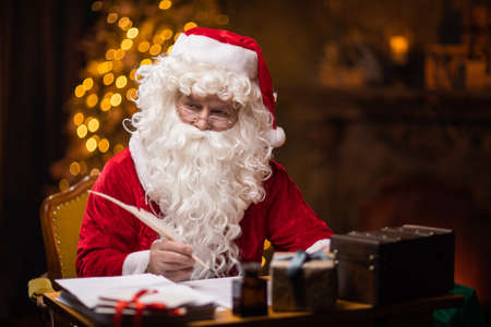 Workplace of Santa Claus. Cheerful Santa is writing the letter while sitting at the table. Fireplace and Christmas Tree in the background. Christmas concept.の写真素材