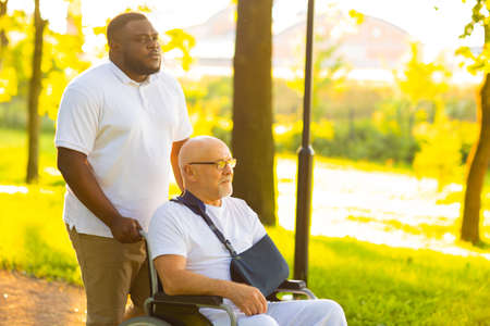 Caregiver and old man in a wheelchair. Professional nurse and patient walking outdoor in the park at sunset. Assistance, rehabilitation and health care.の写真素材