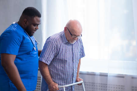 Caregiver is teaching old man to walk with walker. Professional nurse and patient in a nursing home. Assistance, rehabilitation and health.の写真素材