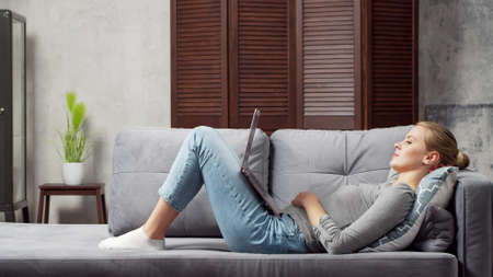 Young woman works using a laptop lying on the couch. Student, entrepreneur or freelancer girl working or studying remotely via internet. Distance learning and job concept.の写真素材