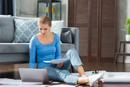 Young woman works with documents using a laptop while sitting on the floor at home. Student,...