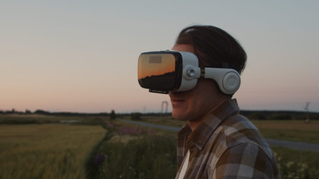 Farmer in virtual reality helmet in front of a sunset agricultural landscape. Man in a countryside...