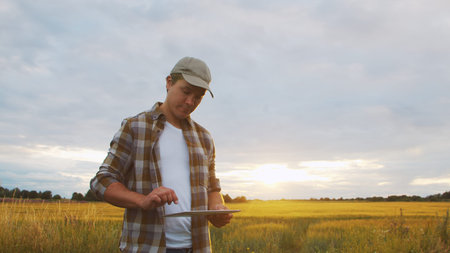Farmer with a tablet computer in front of a sunset agricultural landscape. Man in a countryside...
