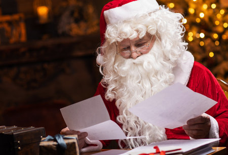 Workplace of Santa Claus. Cheerful Santa is reading letters from children while sitting at the table. Fireplace and Christmas Tree in the background. Christmas concept.の写真素材