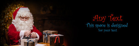 Workplace of Santa Claus. Cheerful Santa is wrapping the gift while sitting at the table. Fireplace and Christmas Tree in the background. Christmas concept.の写真素材