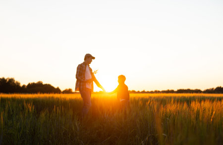 Farmer and his son in front of a sunset agricultural landscape. Man and a boy in a countryside field. Fatherhood, country life, farming and country lifestyle concept.の写真素材