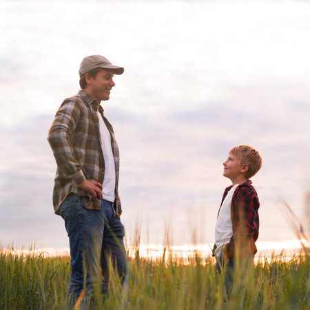 Farmer and his son in front of a sunset agricultural landscape. Man and a boy in a countryside field. Fatherhood, country life, farming and country lifestyle concept.の写真素材