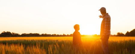 Farmer and his son in front of a sunset agricultural landscape. Man and a boy in a countryside field. Fatherhood, country life, farming and country lifestyle concept.の写真素材
