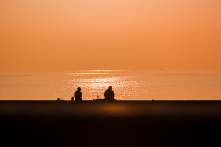 Silhouette of two people sitting on the beach at sunset.の写真素材