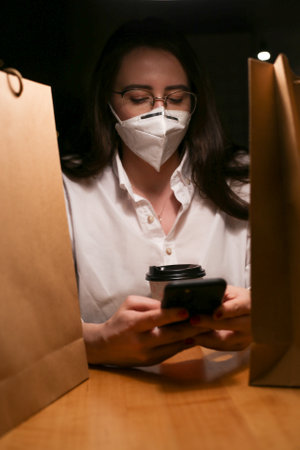 a young woman wears a mask, drinks coffee and looks at the phone writes messagesの写真素材