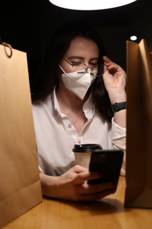 a young woman wears a mask, drinks coffee and looks at the phone writes messagesの写真素材