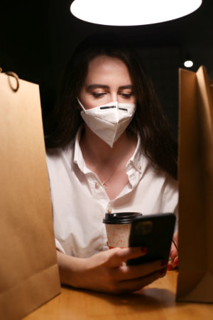 a young woman wears a mask, drinks coffee and looks at the phone writes messagesの写真素材