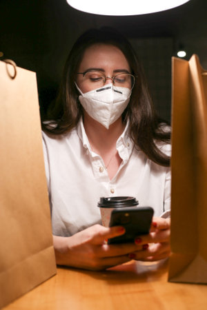 a young woman wears a mask, drinks coffee and looks at the phone writes messagesの写真素材
