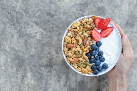 Holding bowl of homemade granola with yogurt and fresh berries on bright gray concrete background from top view.の写真素材