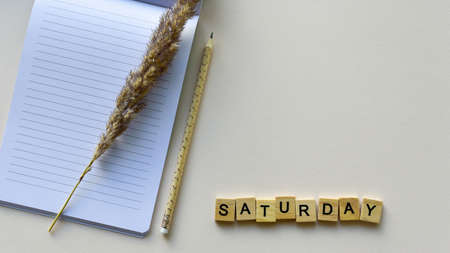 Notepad with pencil and wooden word "Saturday" on a beige paper background. Top view. Close-up. Flat lay.の写真素材